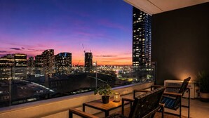 Un balcon avec vue sur la ville au crépuscule, comprenant une table avec des chaises, une plante en pot et un appareil de climatisation.