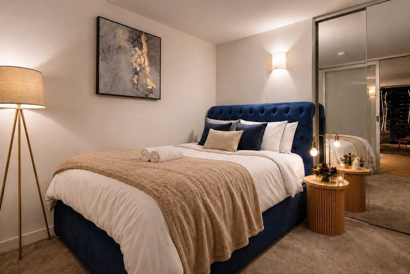 A modern bedroom with a tufted headboard, a large abstract painting, a floor lamp, and a bedside table with a mirror and decorative items.