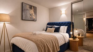 Une chambre à coucher moderne avec une tête de lit capitonnée, un grand tableau abstrait, une lampe sur pied et une table de chevet agrémentée d’un miroir et d’objets décoratifs.
