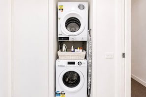 A compact, wall-mounted washer and dryer unit with a control panel and a basket of laundry supplies. - CBD Apartment | Cozy Outdoor Balcony Setup, Heated Pool, Gym, Sauna and Dining (Melbourne)