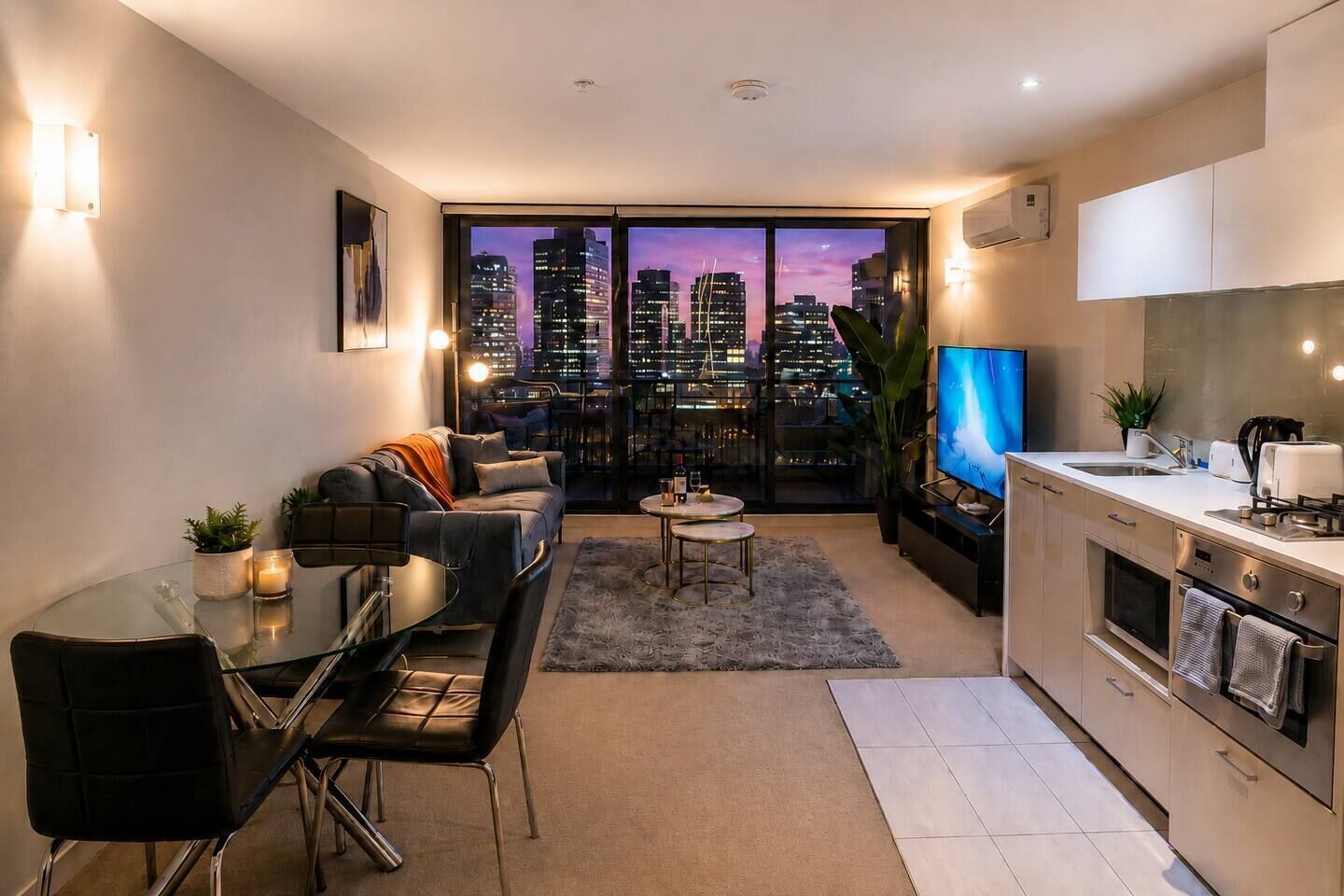 A modern apartment with a dining area, a sofa set, a kitchen, and a large window with a city view.