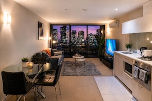 A modern apartment with a dining area, a sofa set, a kitchen, and a large window with a city view. - CBD Apartment | Cozy Outdoor Balcony Setup, Heated Pool, Gym, Sauna and Dining (Melbourne)