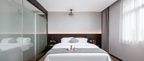 A modern hotel bedroom with a large bed, bedside tables, a nightstand, a glass partition, and a ceiling projector.