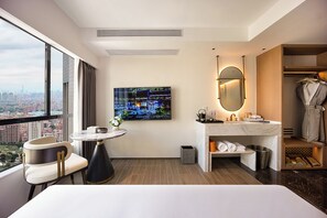 Hotellihuone, jossa on suuri ikkuna, taulu-tv, pyöreä pöytä, moderni sänky, peilillä varustettu kylpyhuone ja erillinen vaatehuone.