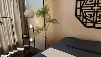 Habitación de hotel con cama, cortinas, un adorno geométrico en la pared, una mesita con lámpara y una planta en maceta.