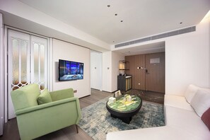 Habitación de hotel moderna con sofá verde, sofá blanco, televisor de pantalla plana, una mesita con objetos decorativos y un armario de madera.