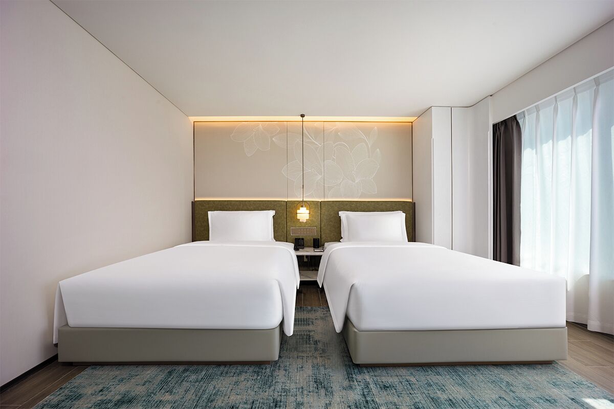 Habitación de hotel con dos camas, un ventanal con cortinas, una lámpara fijada a la pared y una alfombra con textura.