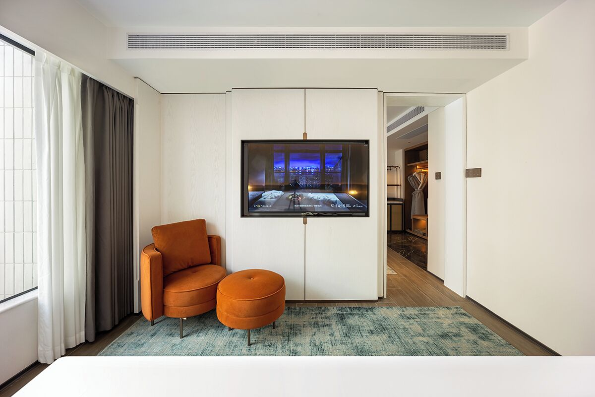 A hotel room with a large flat-screen TV mounted on the wall, a sofa, an ottoman, and a view of the cityscape through the window.