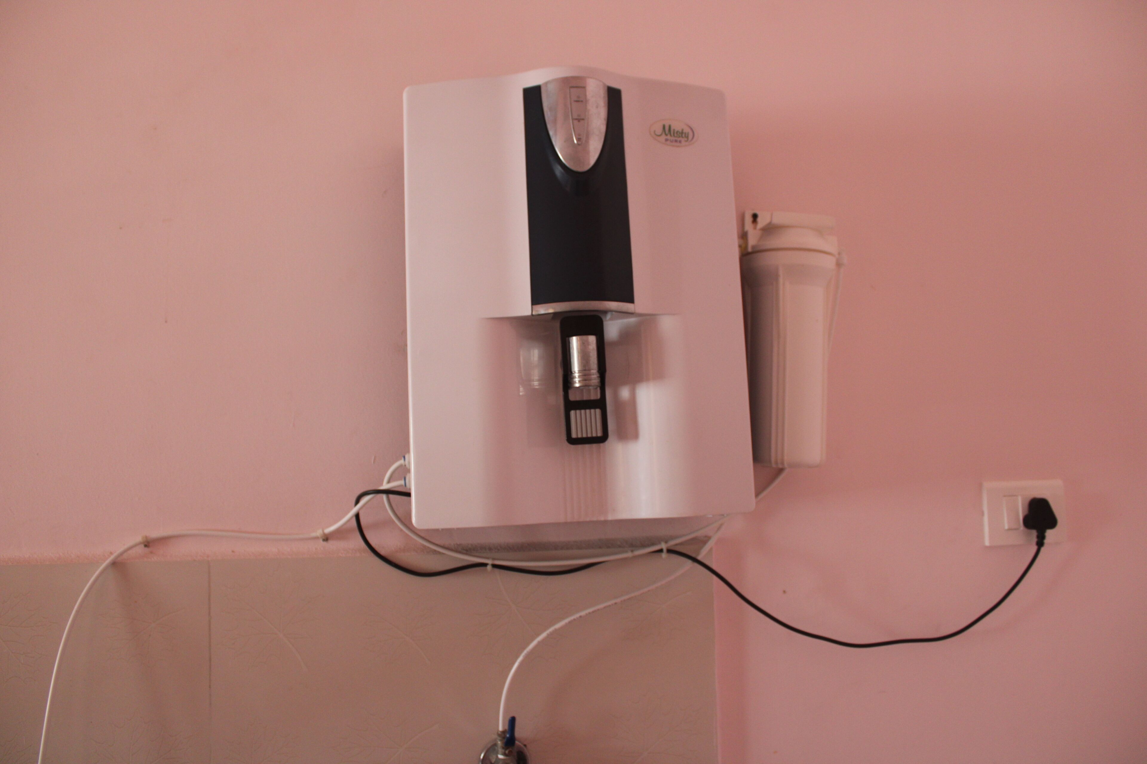A water dispenser mounted on a wall with a power cord plugged into a nearby outlet.