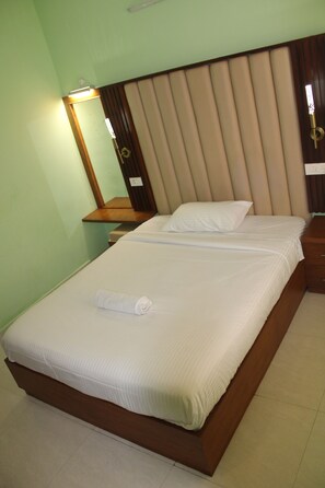 A neatly made bed with a white duvet and pillows, a wooden headboard, a bedside table with a lamp, and a mirror on the wall. - Ramana Palace (Tiruvannamalai)