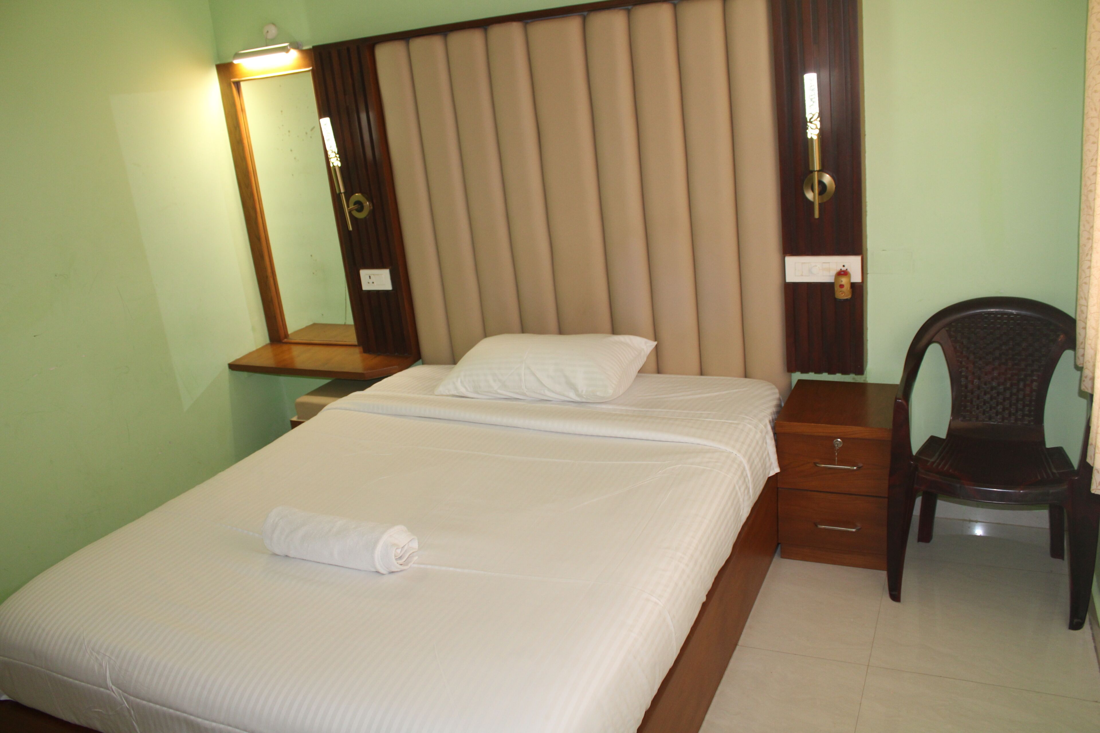 A neatly made bed with a white comforter and pillows, a wooden nightstand with a lamp, and a chair with a patterned backrest.
