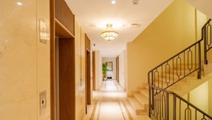A hotel corridor with a staircase on the right, a chandelier, and a plant in the distance. - AKS Palace (Varanasi)