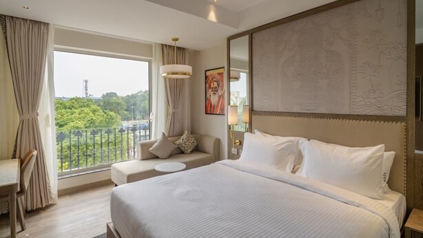 A hotel room with a large bed, a sofa, a desk, and a view of greenery outside. - AKS Palace (Varanasi)