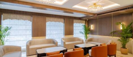 A modern lounge area with sofas, armchairs, and a coffee table, featuring a chandelier and large windows.