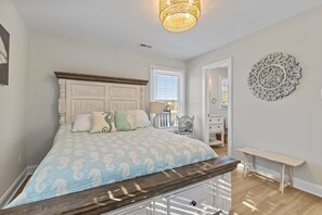 A bedroom with a bed, a bedside table, a bench, a decorative wall piece, and a window with blinds. - SeaEsta | Private Pool + 5 Min Walk to Beach (Emerald Isle)