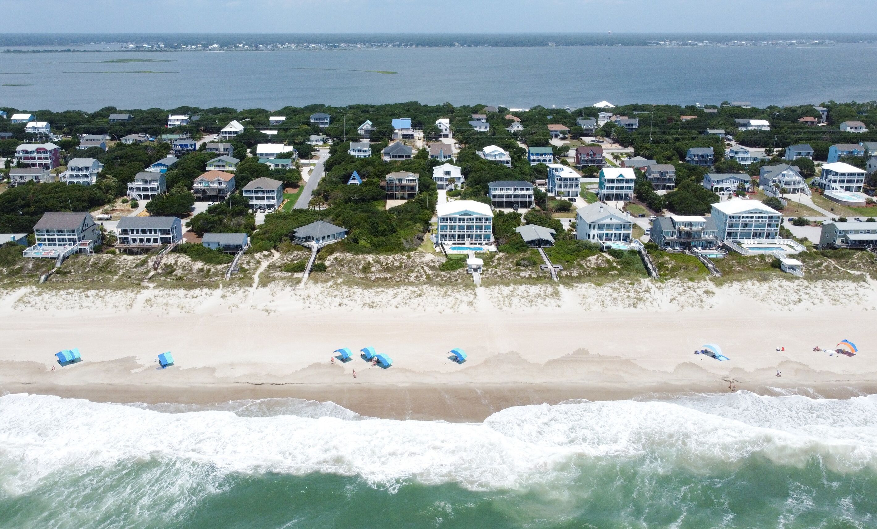 Un quartier résidentiel situé sur la côte, avec des propriétés en bord de mer, notamment des maisons offrant une vue sur l’océan et une plage de sable dotée de parasols.