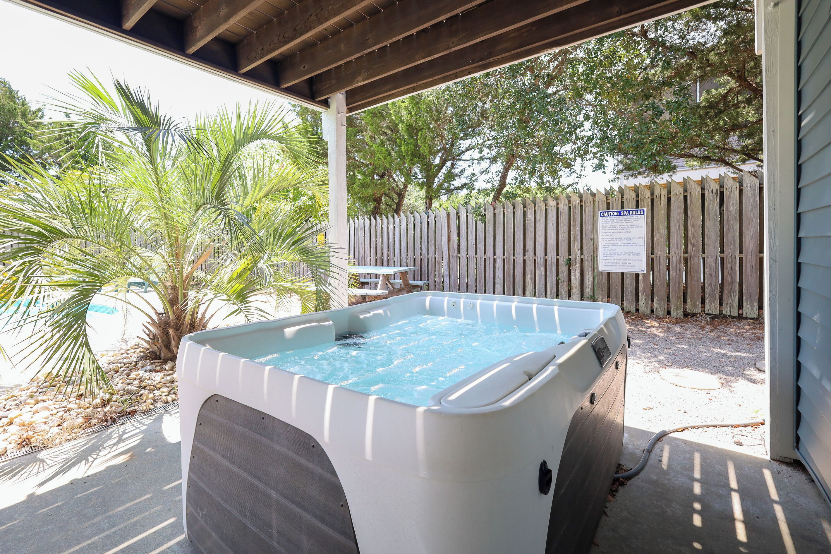 A hot tub in a covered outdoor area with a wooden fence and palm trees in the background.