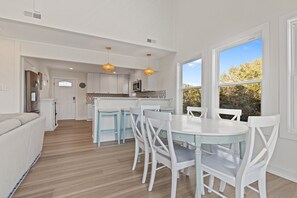 A modern kitchen with white cabinetry, a dining area with a white table and chairs, and a view of the outdoors through large windows. - SeaEsta | Private Pool + 5 Min Walk to Beach (Emerald Isle)