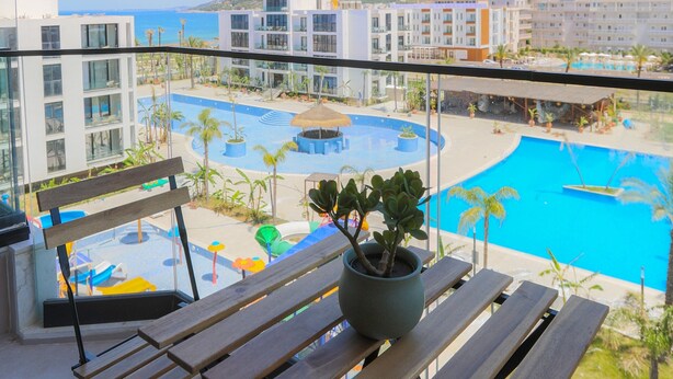 Un balcón con un banco de madera y una planta en maceta, con vista a una piscina y edificios modernos.