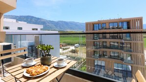 A balcony with a table set for two, a potted plant, and a view of a modern building and mountains. - Modern Pool Side Studio Palm (Orikum)