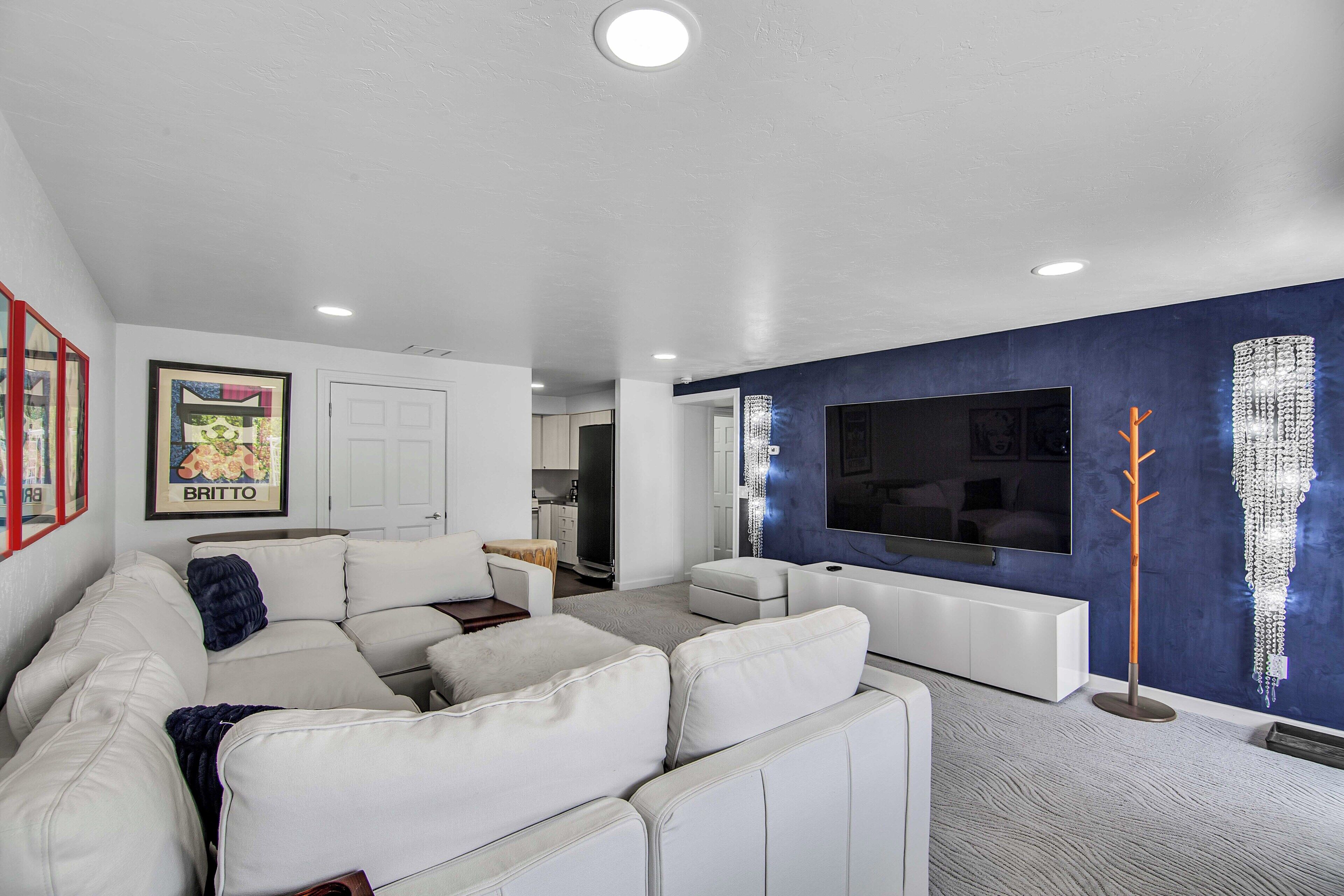 Un salon moderne avec un canapé d’angle blanc, un téléviseur à écran plat fixé sur un mur bleu qui sert de point de couleur, et une petite cuisine en arrière-plan.