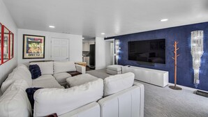A modern living room with a white sectional sofa, a flat-screen TV mounted on a blue accent wall, and a small kitchen in the background.