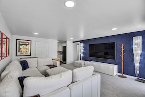 A modern living room with a white sectional sofa, a flat-screen TV mounted on a blue accent wall, and a small kitchen in the background.