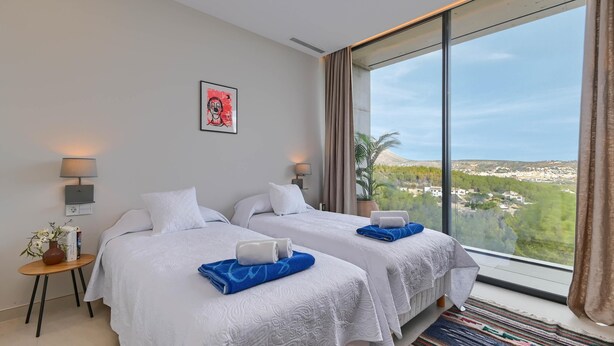 Camera d'albergo con due letti, un'ampia finestra vista città, un piccolo tavolo con una lampada e un quadro appeso alla parete.