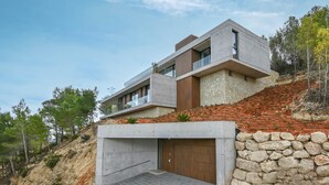 Una moderna casa a due piani con un muro di sostegno in pietra, un garage e uno sfondo collinare.