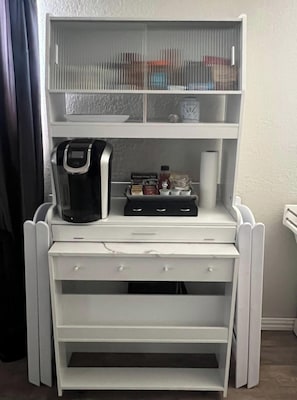 Um minibár branco com máquina de café, sachês de condimentos e uma garrafa de água.