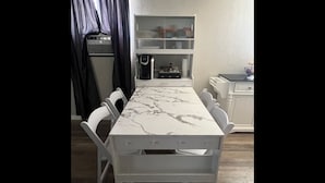 Una mesa de comedor con superficie de mármol, rodeada de sillas blancas, en una habitación con un armario blanco y una cafetera sobre la encimera.