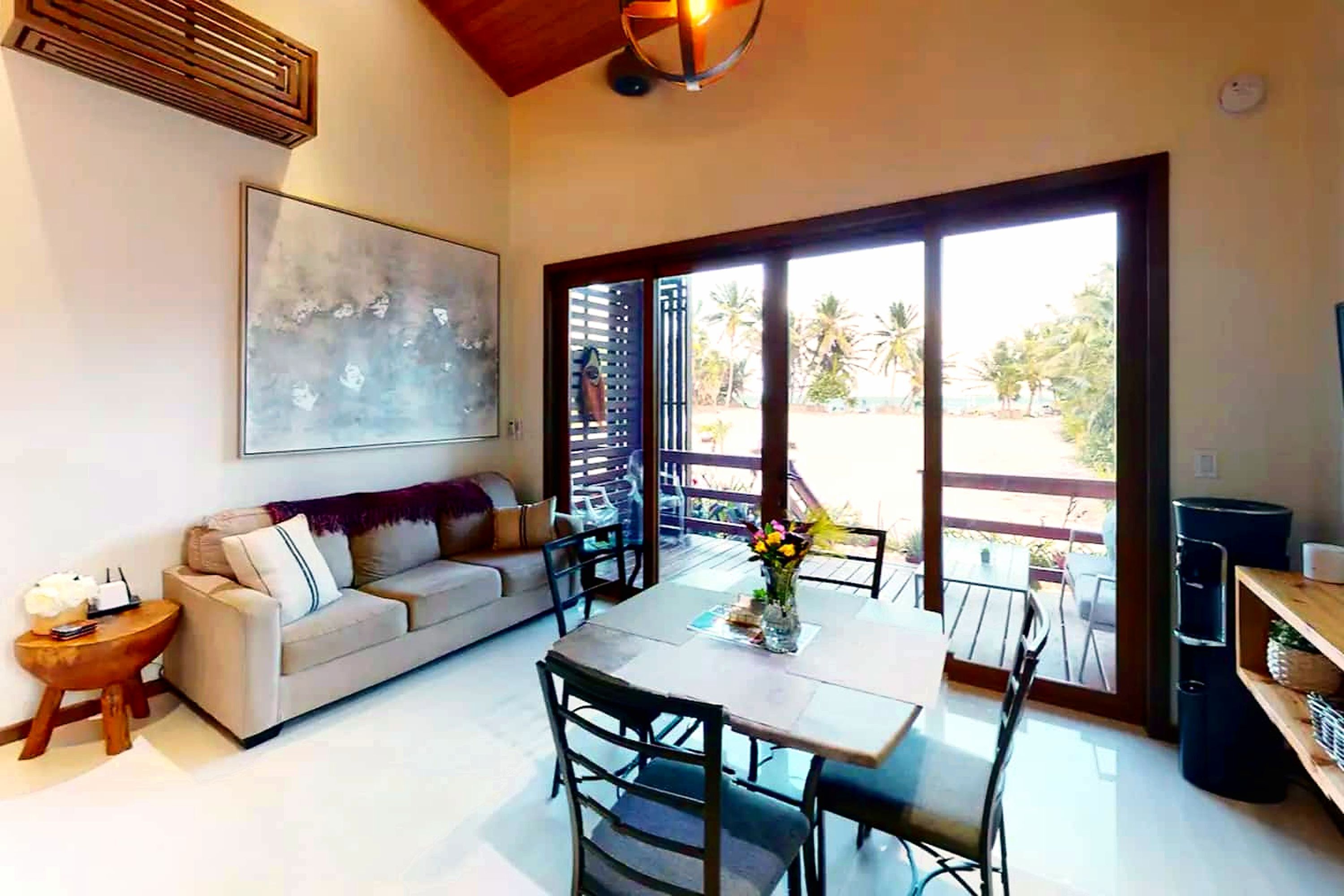 Un'ampia zona giorno con divano, tavolo da pranzo e sedie, un'ampia finestra con vista su palme e una ventola a soffitto.
