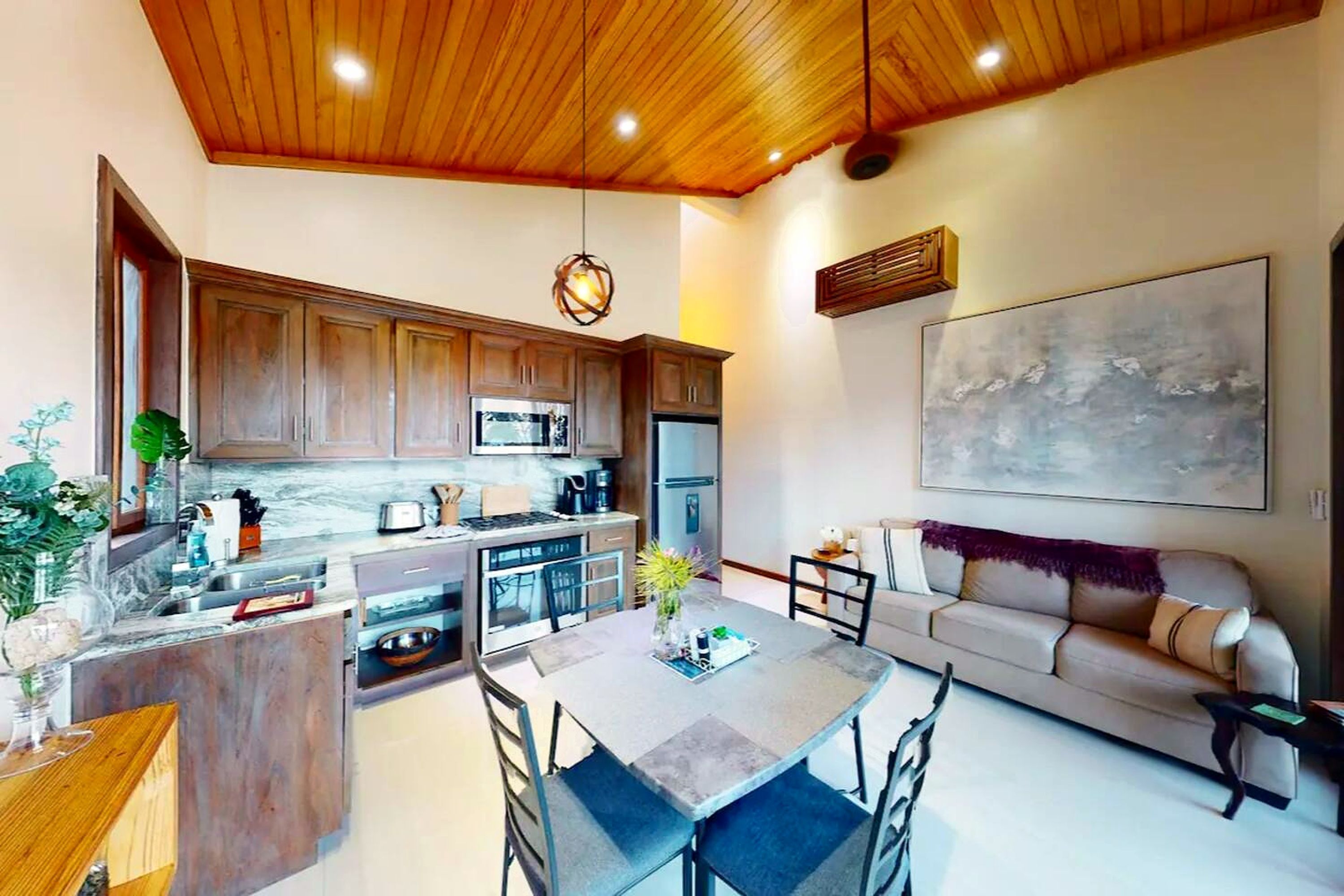 A modern kitchen with wooden cabinets, a dining area with a table and chairs, and a living room with a sofa and a large abstract painting.