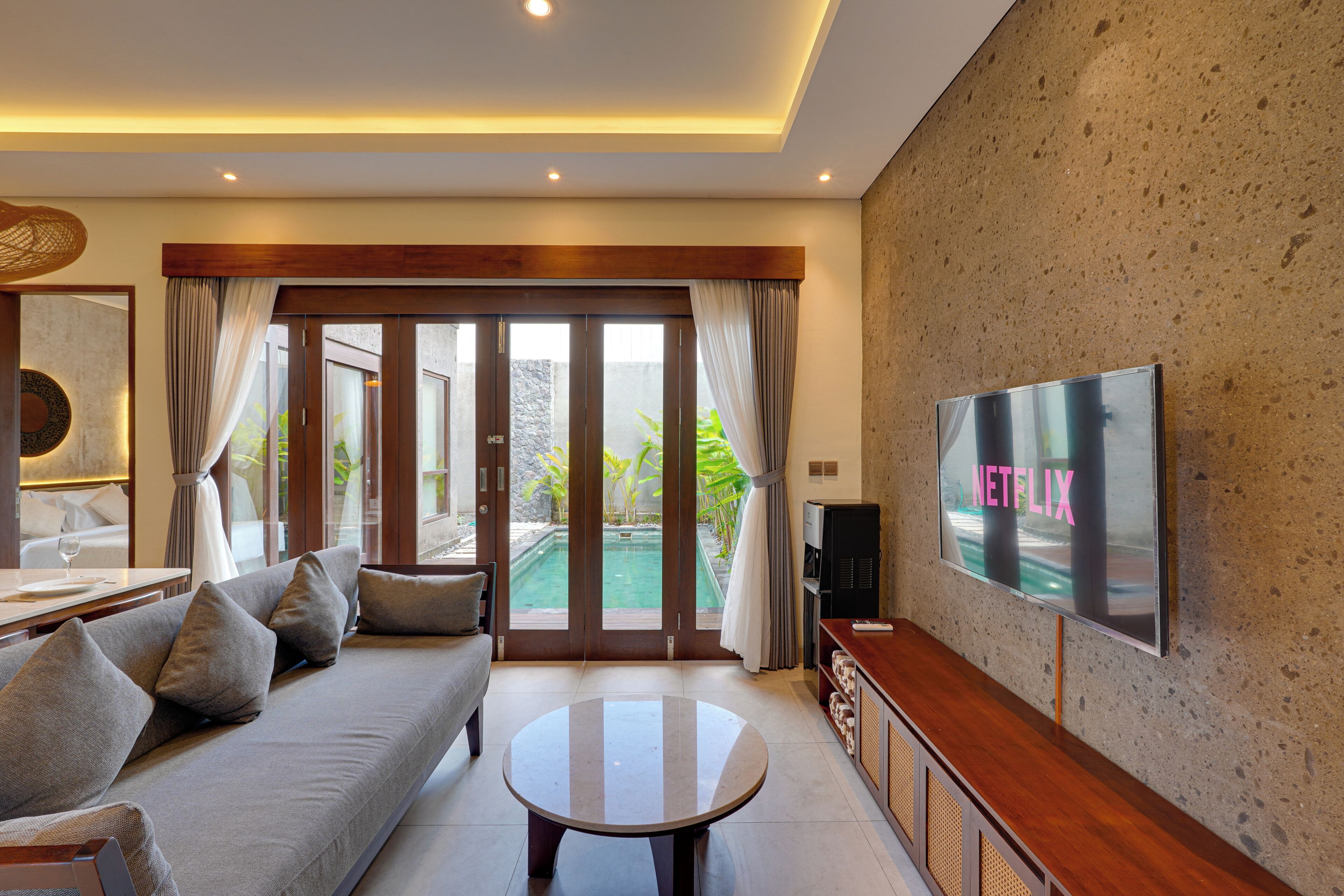 A modern living room with a sofa, a coffee table, a flat-screen TV, and a sliding glass door leading to a pool area.