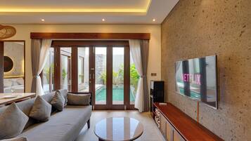 A modern living room with a sofa, a coffee table, a flat-screen TV, and a sliding glass door leading to a pool area.