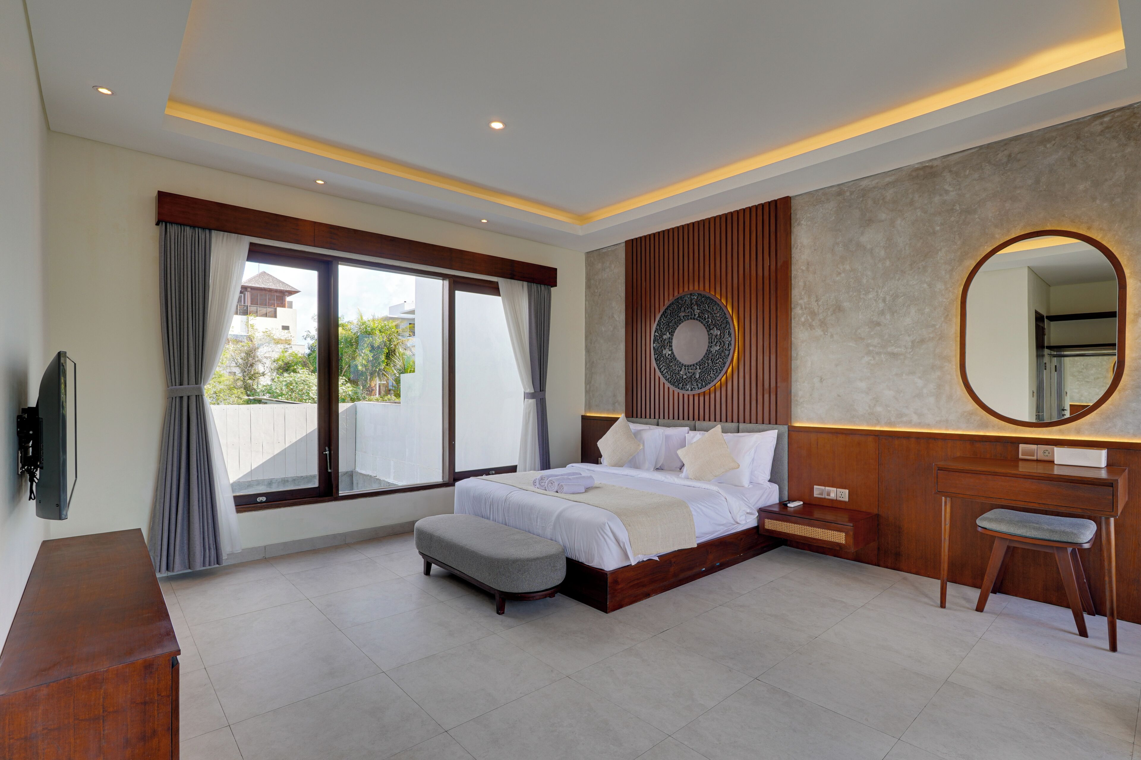 A modern bedroom with a large bed, a wooden dresser, a vanity with a mirror, and a television on the wall.