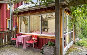 A covered outdoor seating area with a table and chairs, a wooden deck, and a window overlooking a garden. - Cozy home in Ellös with sauna (Ellös)