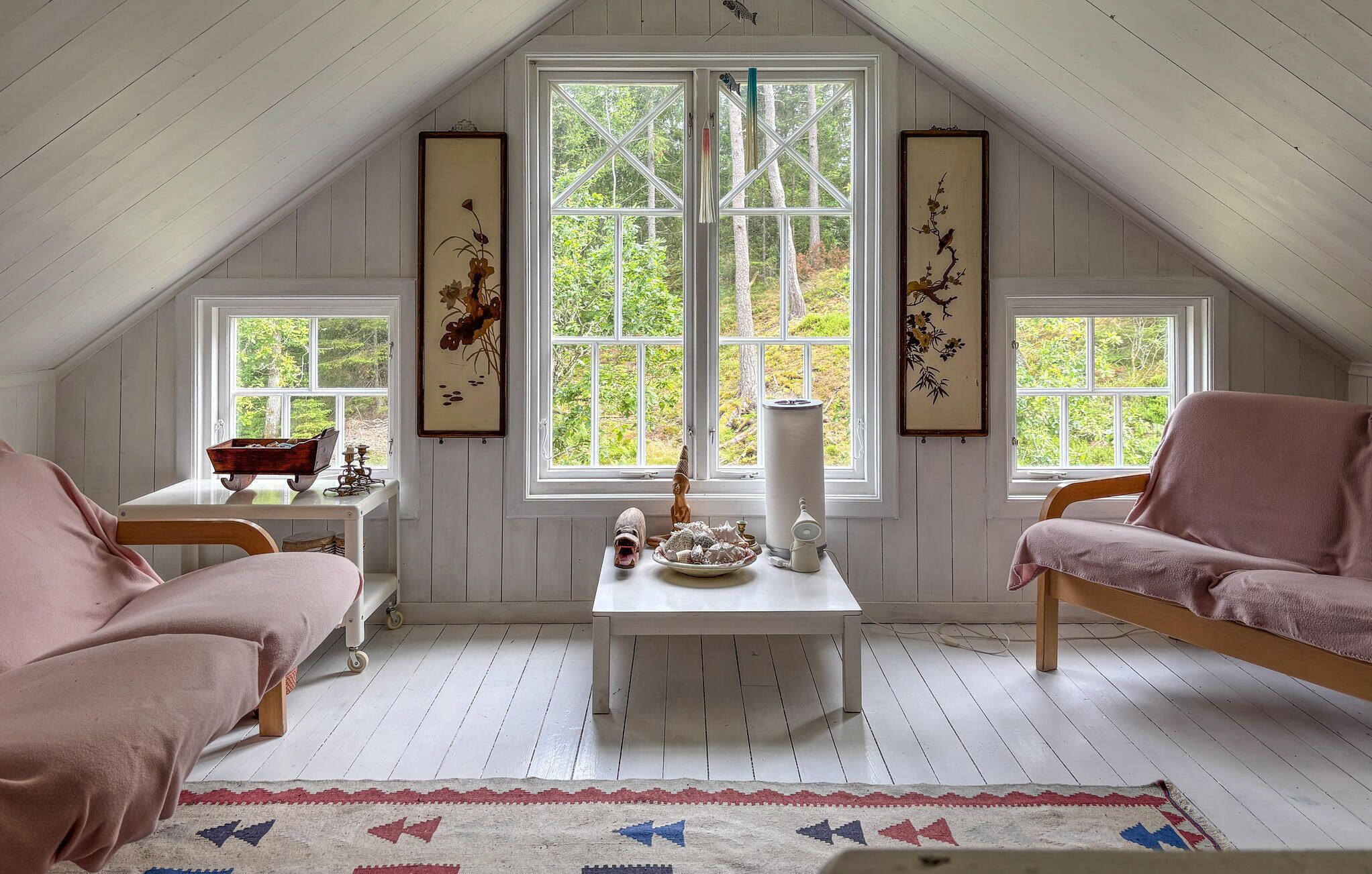 A cozy attic room with a sloped ceiling, white wooden walls, and large windows offering a view of greenery outside.