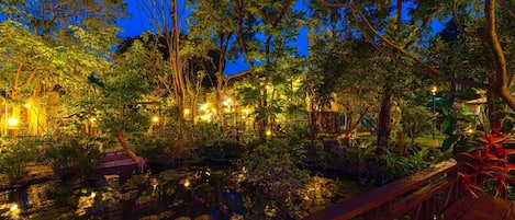Un jardín nocturno con un estanque, árboles y un puente de madera.