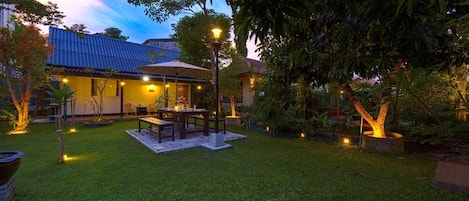 Un patio al aire libre bien iluminado con una mesa y sillas para comer, rodeado de abundante vegetación y una casa con techo azul.