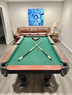 A pool table with two cue sticks crossed in the center, a brown leather sofa, and a blue abstract painting on the wall.
