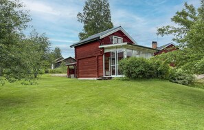 A red wooden house with a glass door entrance, surrounded by a well-maintained lawn and dense greenery. - 1 bedroom lovely home in Leksand (Leksand)