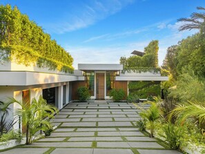 A modern house with a central entrance, surrounded by a garden with various plants and a paved walkway.