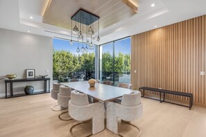 A modern dining area with a large table, six chairs, and a bench. There is a wooden bench along the wall, a black console table with decorative items, and a view of trees through large windows.