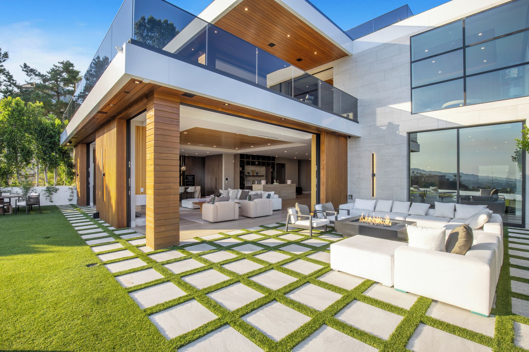Uma casa moderna de dois andares com uma ampla área de estar ao ar livre, incluindo uma lareira, sofás brancos e um gramado bem cuidado.