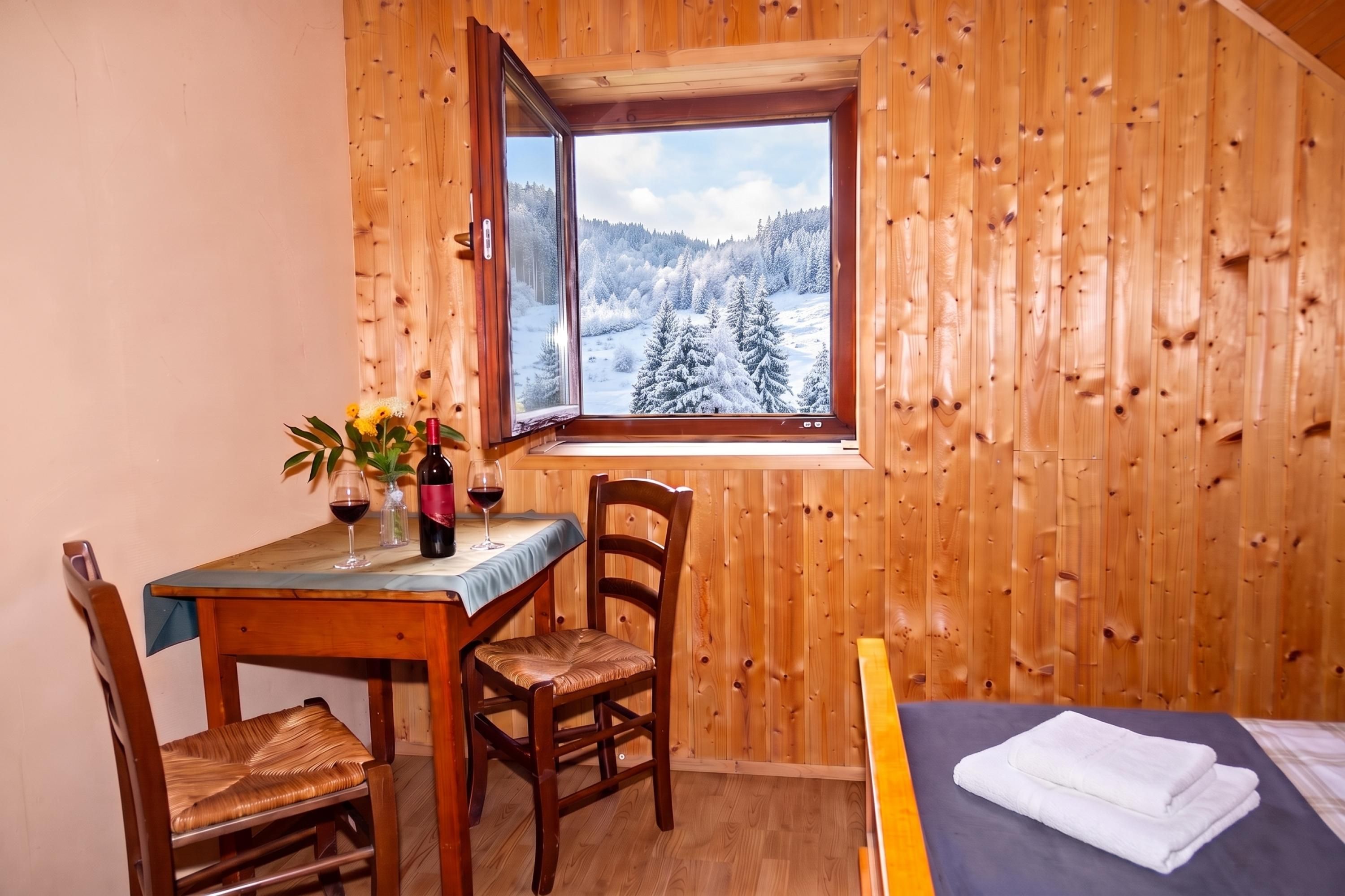 Interior de madera con una mesa de comedor, sillas, una ventana que ofrece vistas a un paisaje nevado y una cama con una toalla blanca doblada.
