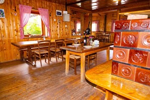 A wooden interior with multiple dining tables and chairs, a kitchen area with a refrigerator, and a window with checkered curtains. - Alpine Lodge Goska Ravan Rooms (Radovljica)