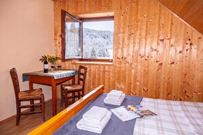 A wooden cabin room with a bed, a table, chairs, and a window with a snowy forest view. - Alpine Lodge Goska Ravan Rooms (Radovljica)