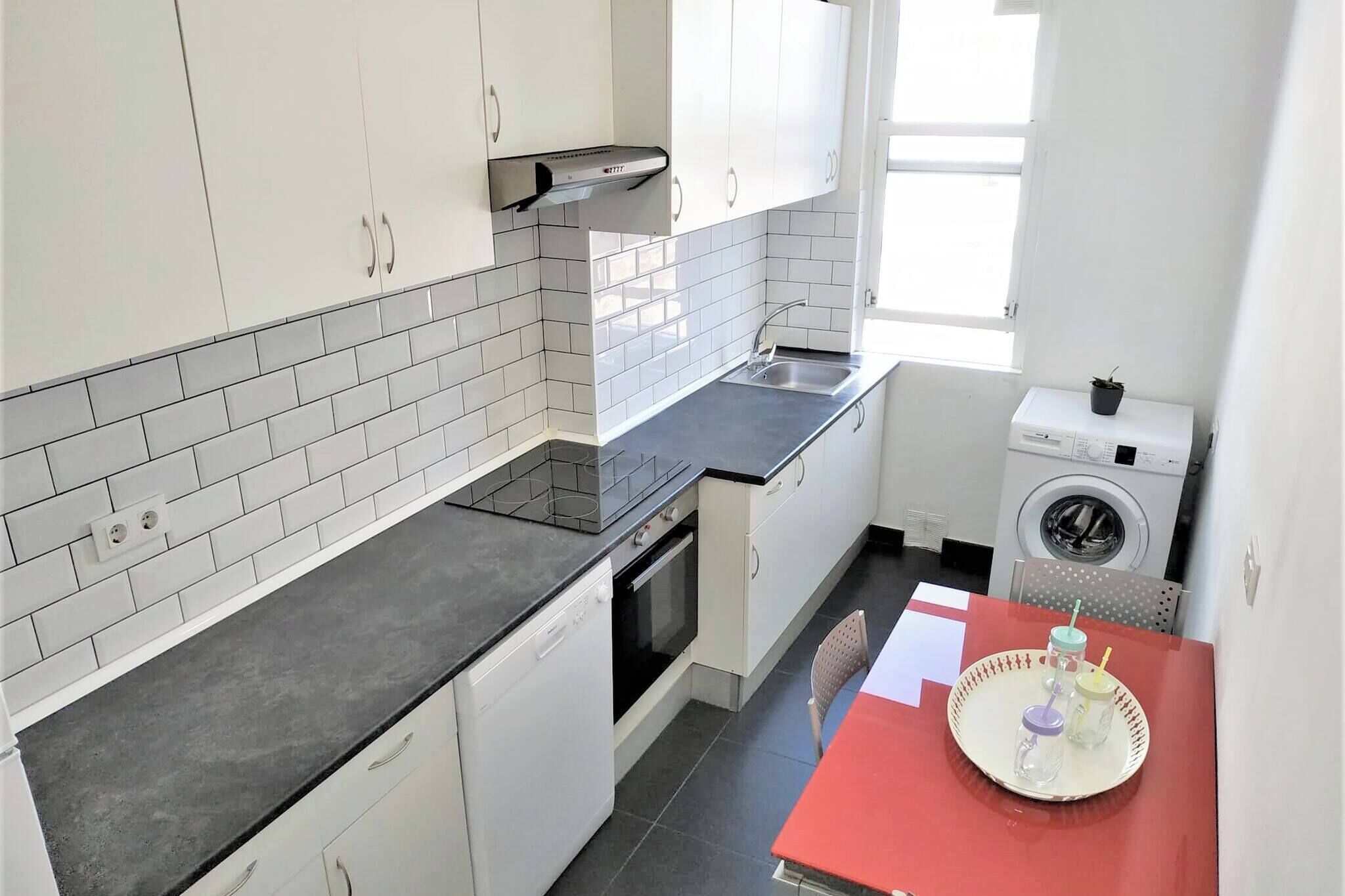 A compact kitchen with white cabinets, a black countertop, a built-in oven, a washing machine, and a red table with a tray.