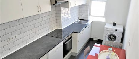 Cocina compacta con armarios blancos, encimera negra, horno empotrado, lavadora y una mesa roja con bandeja.